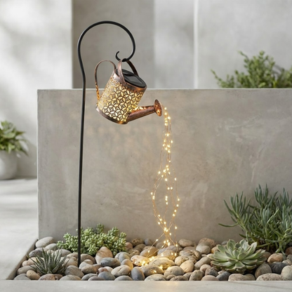 Decorative solar light shaped like a watering can with fairy lights on pebbles near plants.
