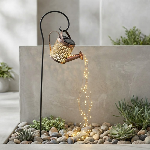 Decorative solar light shaped like a watering can with fairy lights on pebbles near plants.