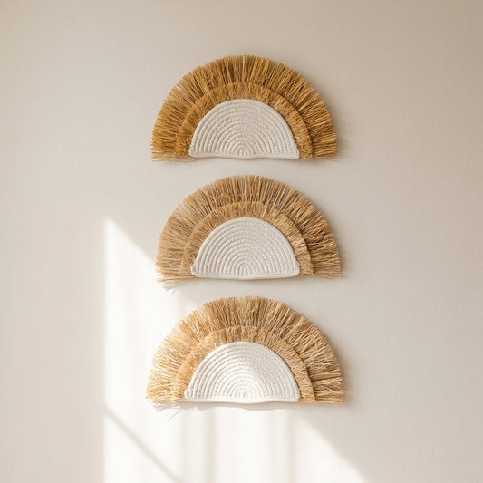 Three decorative wall art pieces with raffia fringes on a plain wall.